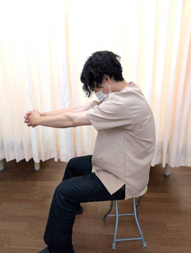 椅子に座り、両腕を前で組んで背中を丸め、肩甲骨まわりを伸ばすストレッチ